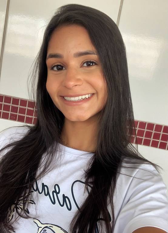 Foto de Camila Santos do Nascimento, uma jovem mulher de pele parda, com cabelos lisos abaixo dos ombros. Seus cabelos e olhos são castanho-escuros. Seu queixo possui covinha. Usa uma camiseta branca com alguns escritos. Está com a cabeça levemente inclinada para a esquerda, sorrindo, e faz a foto no estilo selfie.