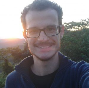 Foto de Felipe Fernando Furlan, um homem de pele branca, com cabelos curtos mais volumosos na parte de cima.  Ele está de óculos de grau com armação preta e quadrada, usa uma camiseta preta, um agasalho azul marinho por cima e sorri.