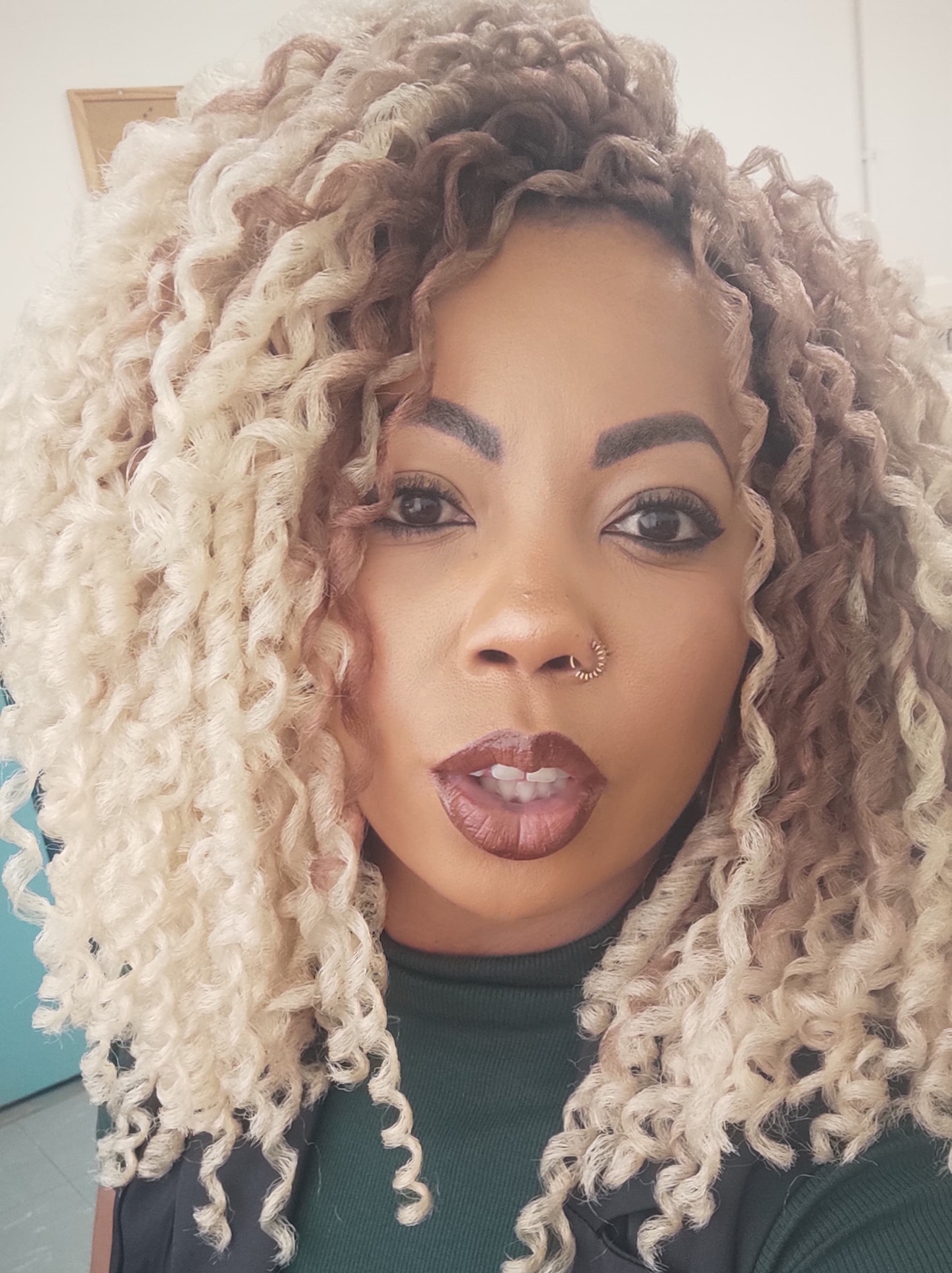 Foto de Raquel Santos Diniz, uma mulher de pele negra, com cabelos bem cacheados na cor loira, Seus olhos são castanho-escuro. Ela usa batom na cor nude. Tem um piercing de argola na narina direita e usa uma blusinha na cor verde escuro e colete preto por cima.