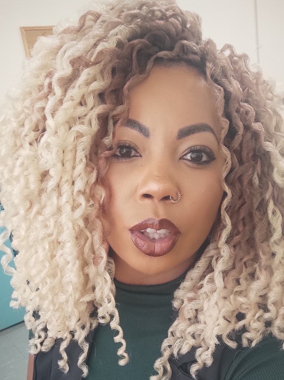 Foto de Raquel Santos Diniz, uma mulher de pele negra, com cabelos bem cacheados na cor loira, Seus olhos são castanho-escuro. Ela usa batom na cor nude. Tem um piercing de argola na narina direita e usa uma blusinha na cor verde escuro e colete preto por cima.