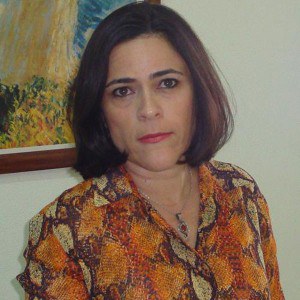 Foto de Mônica Lopes Aguiar, uma mulher de pele branca, com cabelos lisos e curtos, na altura do queixo. Seus cabelos e olhos são castanhos. Ela usa um colar com pingente e uma camisa com estampa em tons de laranja e marrom.