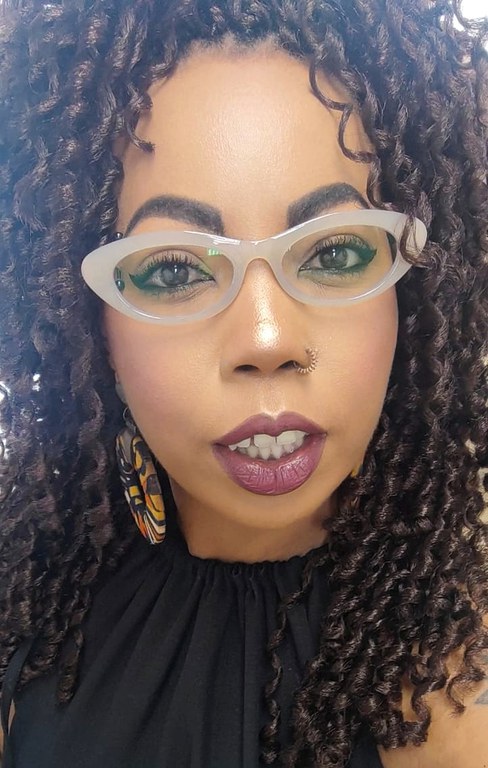 Foto de Raquel Santos Diniz, uma mulher de pele negra, com cabelos cacheados na cor castanho-escura, estilo black power. Seus olhos são castanho-escuro. Ela usa óculos de grau estilo gatinho na cor bege clara e seus lábios estão com batom na cor nude. Ela tem um piercing de argola na narina direita e usa um vestido na cor preta.