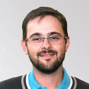 Foto de Rodrigo Béttega, um homem de pele branca, com cabelos lisos e curtos e barba com cavanhaque. Seus cabelos, olhos e barbas são castanhos. Ele está de óculos de grau com armação retangular prateada e usa uma camisa polo com gola azul, branca, preta e cinza. 